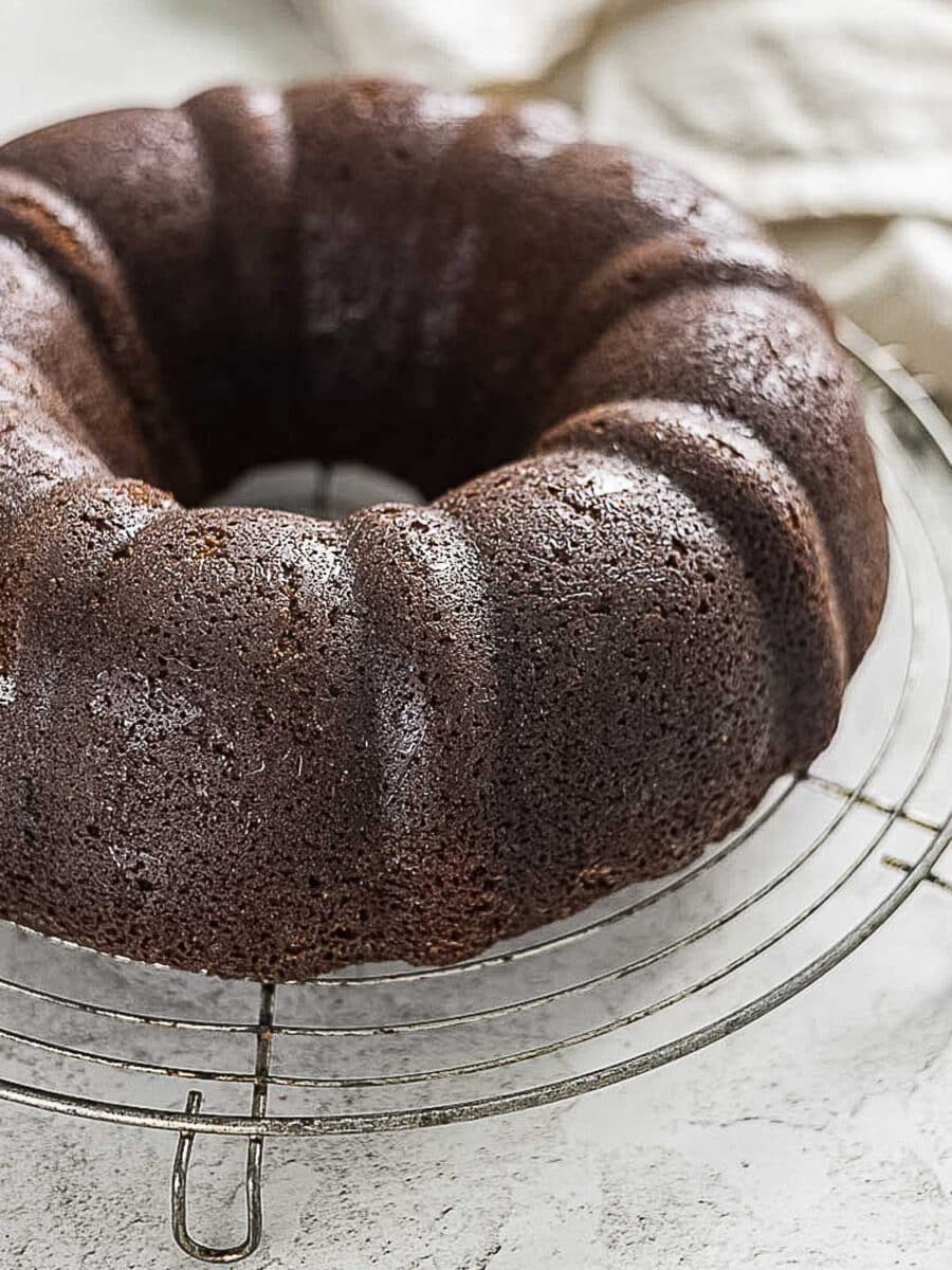 A chocolate bundt cake cools on a round metal wire rack, with a neutral background and a light cloth partially visible.