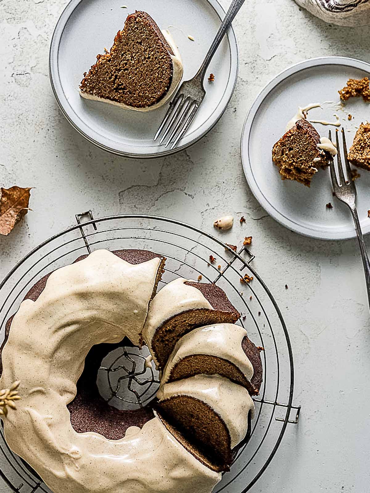 A bundt cake with icing is on a cooling rack, with a few slices cut. Two plates nearby have cake slices, each with a fork and some crumbs.