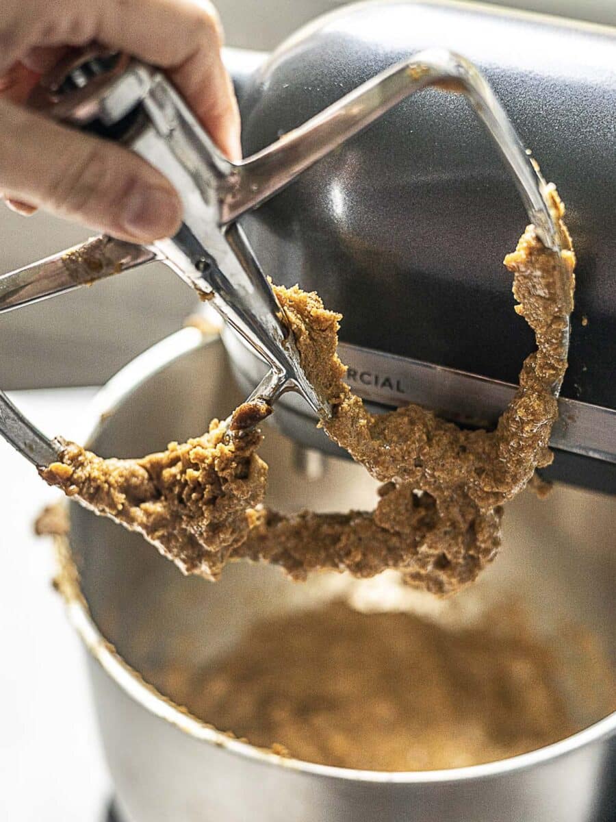 A close-up of a person lifting the paddle attachment from a stand mixer with cookie dough clinging to it.