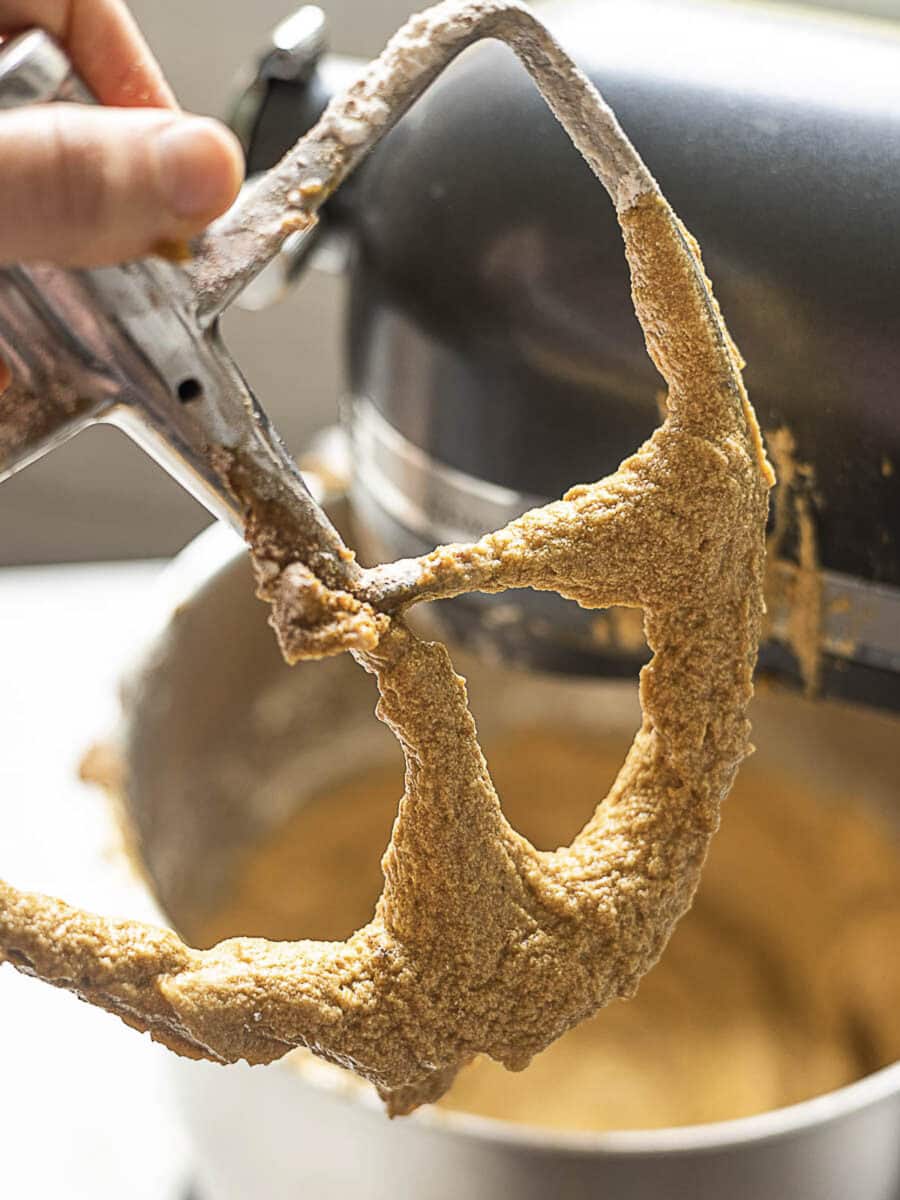A close-up of a stand mixer paddle attachment covered in light brown cookie dough, held above a mixing bowl.