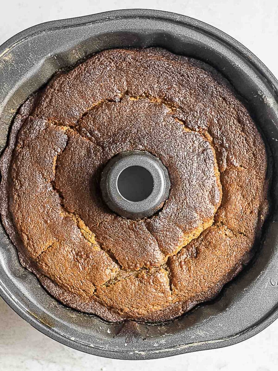 A baked bundt cake with a cracked, golden-brown top sits inside a metal bundt pan on a light surface.