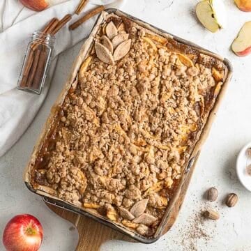 A baked apple crumble tray topped with oat streusel sits on a wooden board, surrounded by fresh apples, cinnamon sticks, and nutmeg on a white surface.