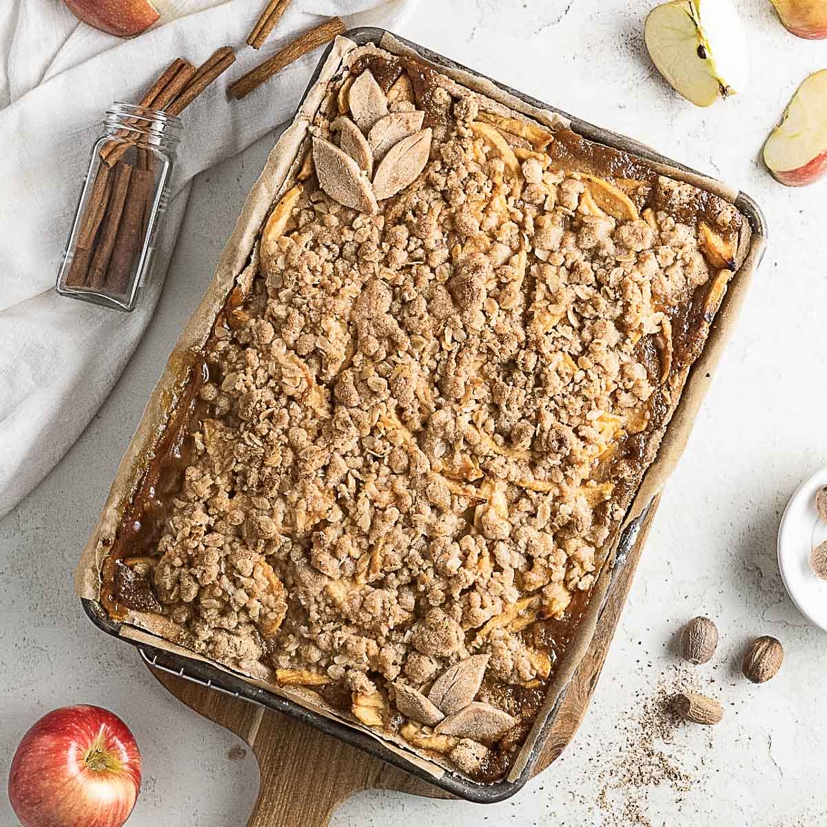 A baked apple crumble tray topped with oat streusel sits on a wooden board, surrounded by fresh apples, cinnamon sticks, and nutmeg on a white surface.