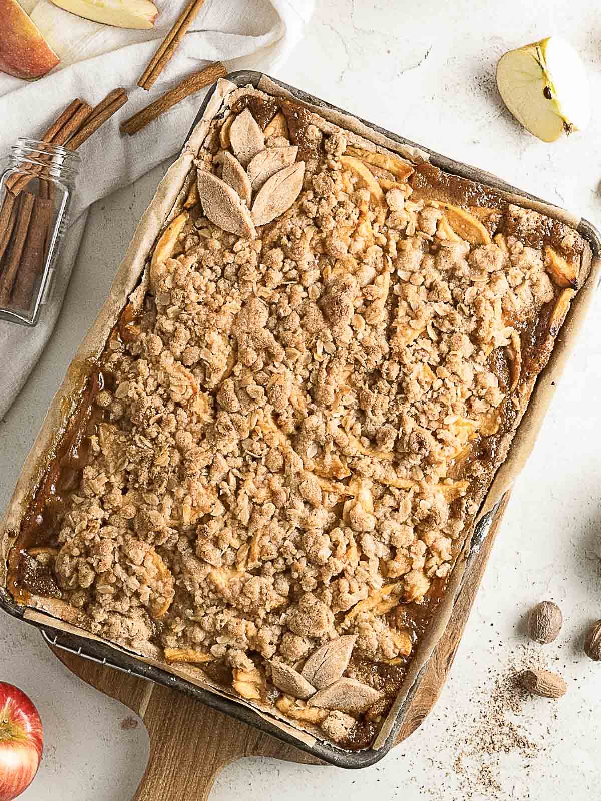 A rectangular apple crumble dessert in a baking dish, topped with streusel and decorative pastry, sits on a white surface with apples and cinnamon sticks nearby.