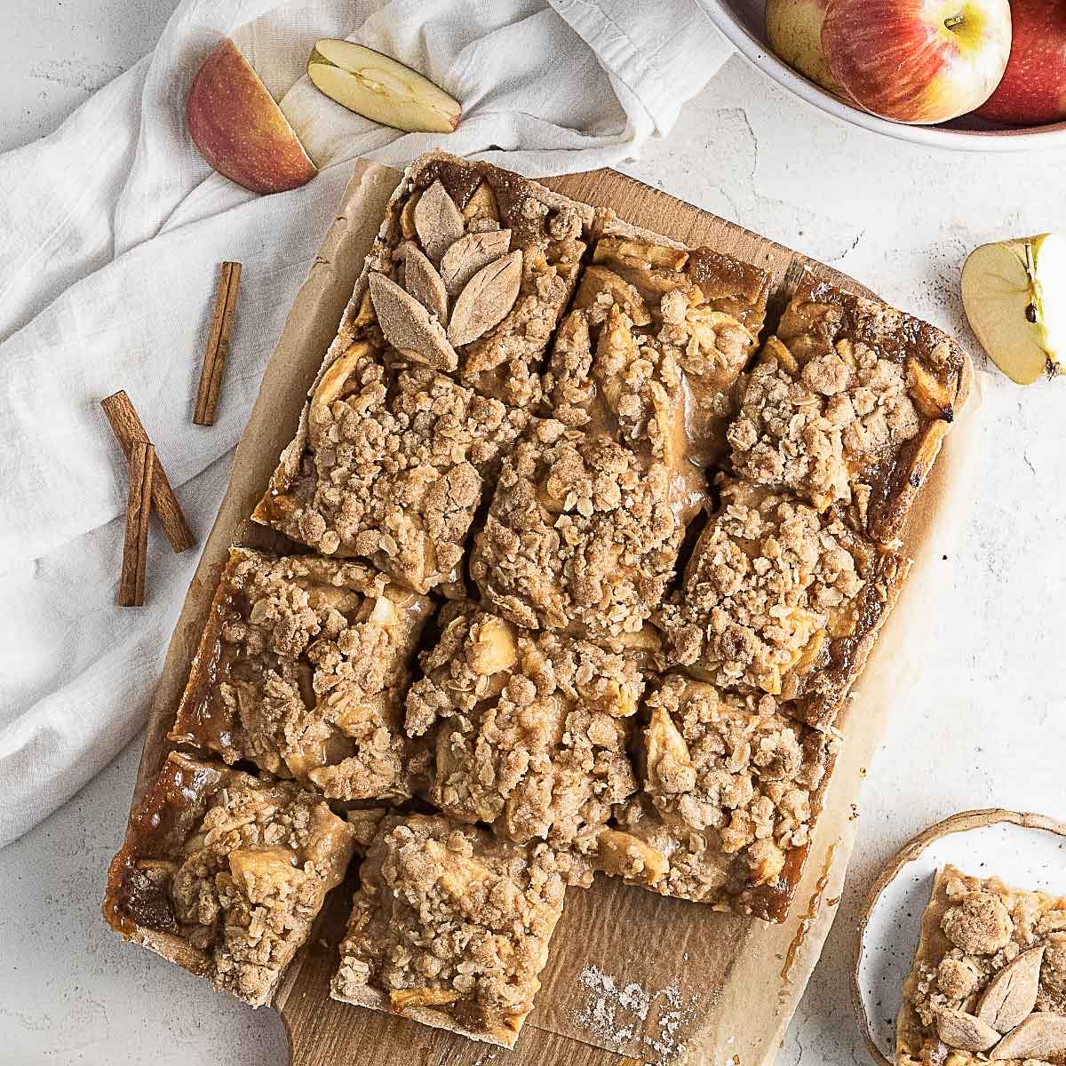 A wooden board with sliced apple crumble bars, surrounded by apple pieces, cinnamon sticks, and a bowl of apples on a light surface.