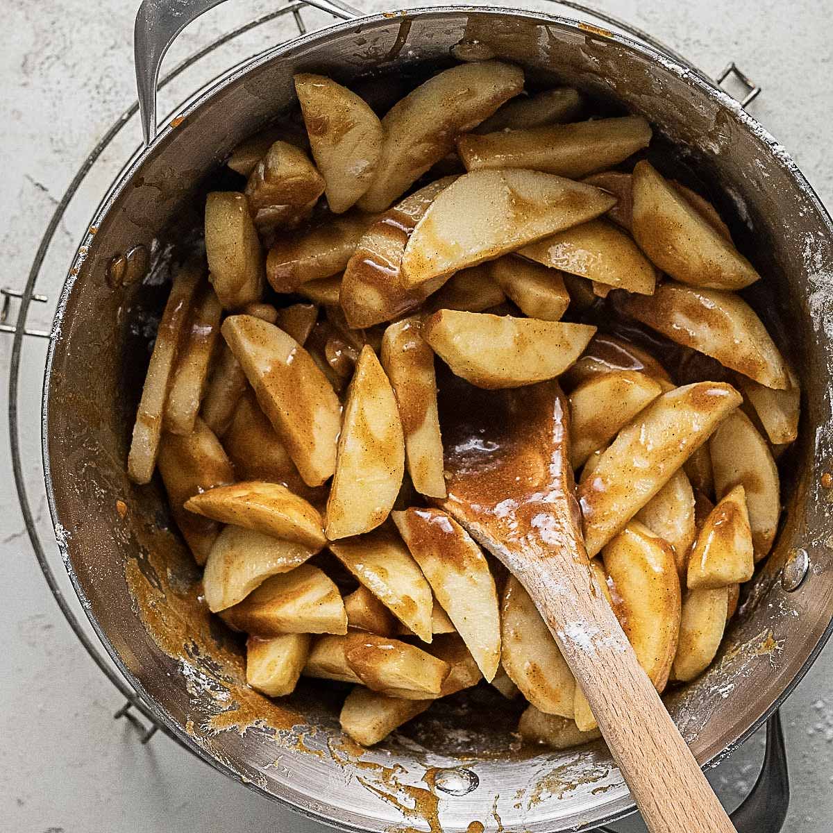 Sliced apples coated in brown cinnamon-sugar mixture being stirred with a wooden spoon in a metal pot.