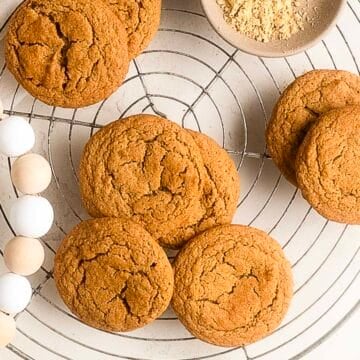 Round ginger cookies arranged on a wire cooling rack, with a small bowl of ground ginger and a wooden bead garland nearby.