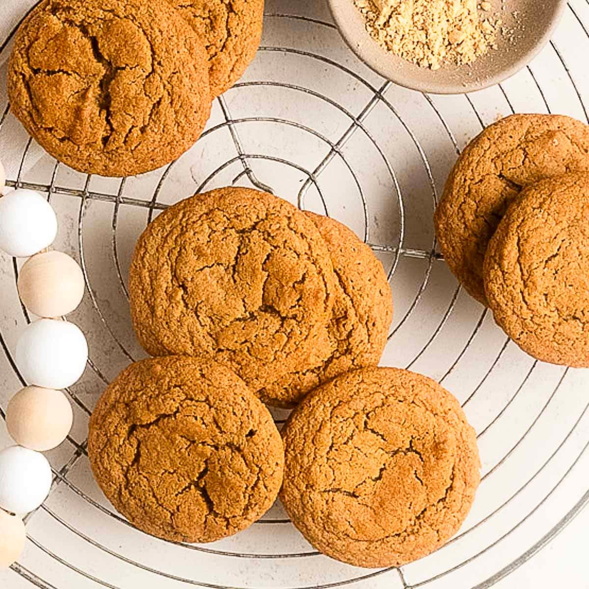 Round ginger cookies arranged on a wire cooling rack, with a small bowl of ground ginger and a wooden bead garland nearby.