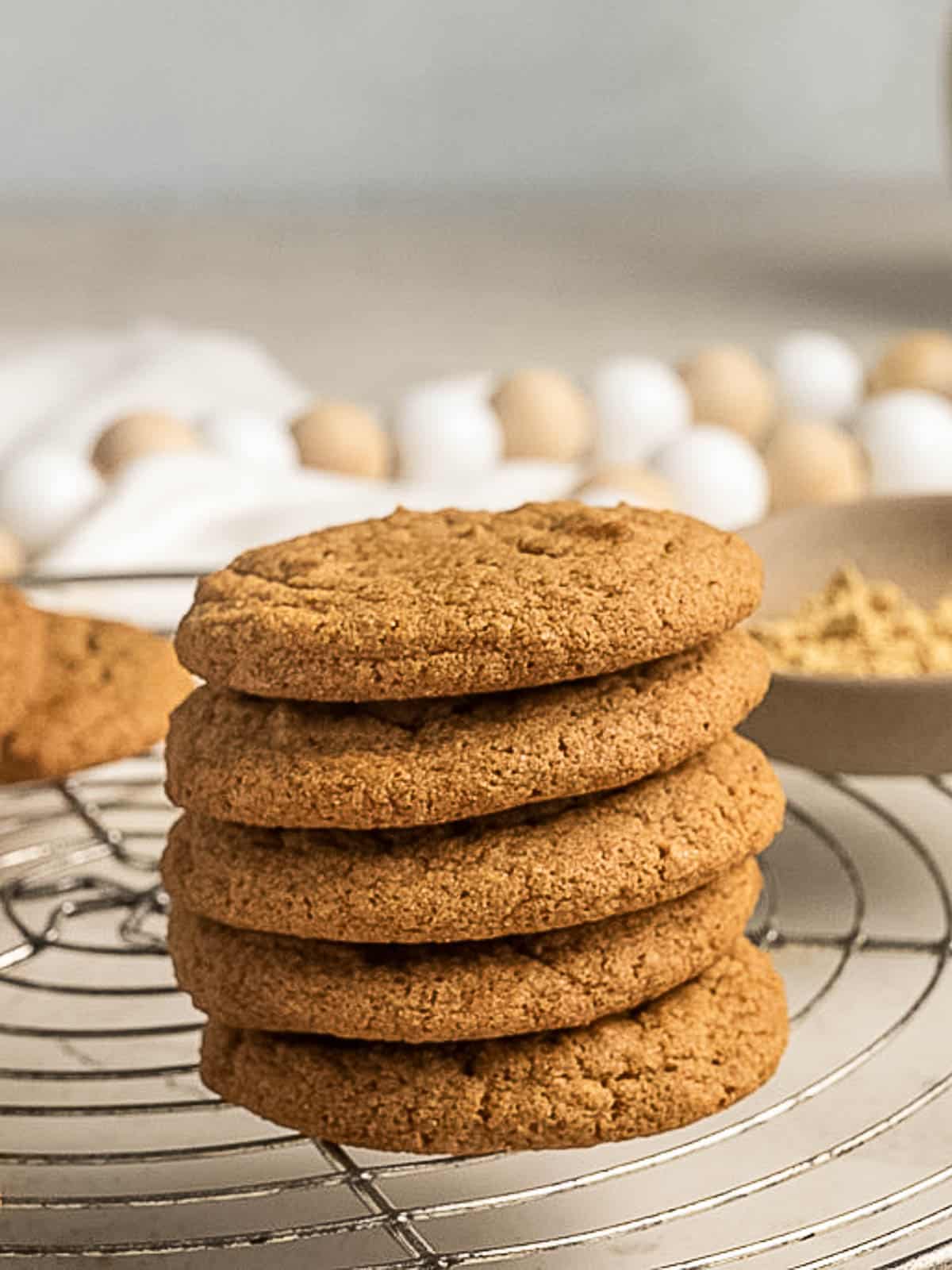 A stack of six golden brown cookies is placed on a round wire cooling rack with eggs visible in the blurred background.