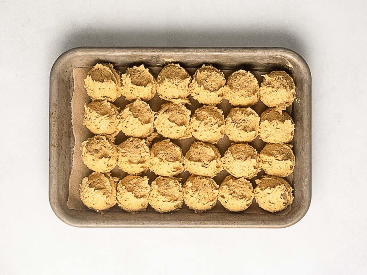 A baking tray lined with parchment paper holds 24 evenly spaced scoops of raw cookie dough on a white surface.