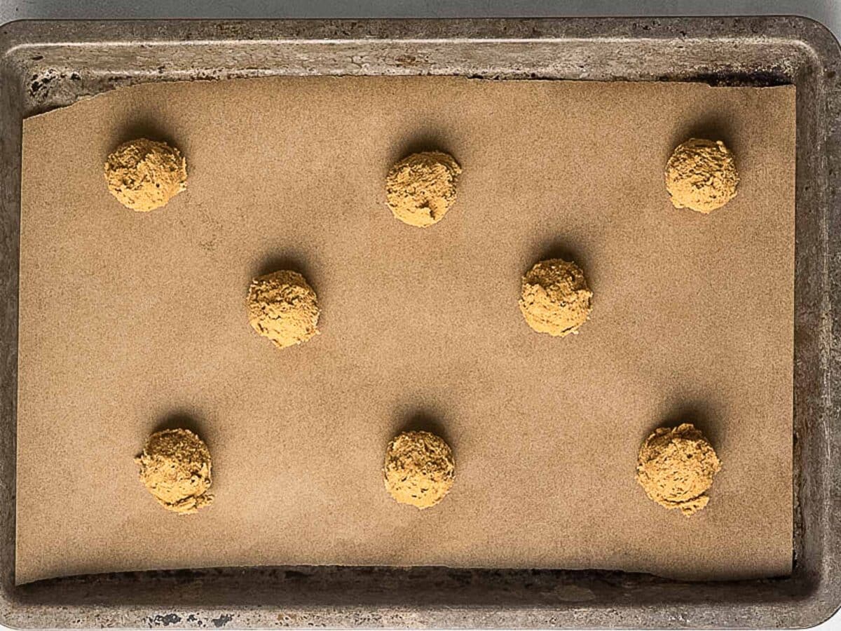 A baking tray lined with parchment paper holds eight evenly spaced scoops of raw cookie dough, ready to be baked.