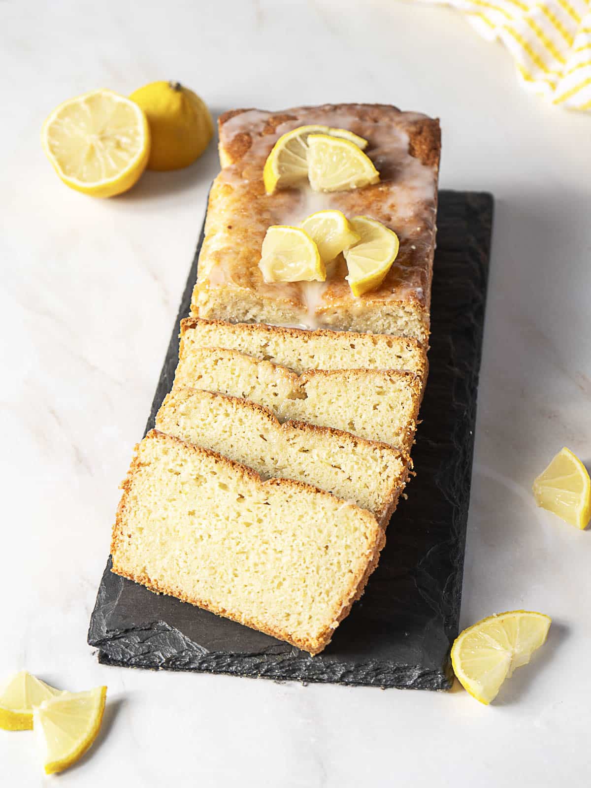 A loaf of lemon bread with a glaze, topped with lemon slices, is partially sliced and displayed on a black slate serving board. Whole and cut lemons are placed nearby.