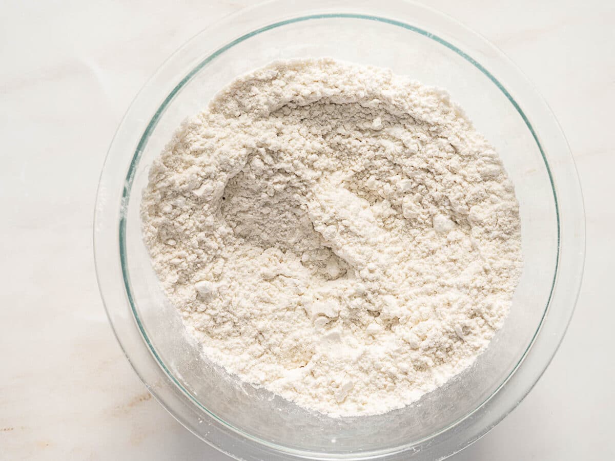 A clear glass bowl filled with a mixture of dry flour on a light-colored surface.