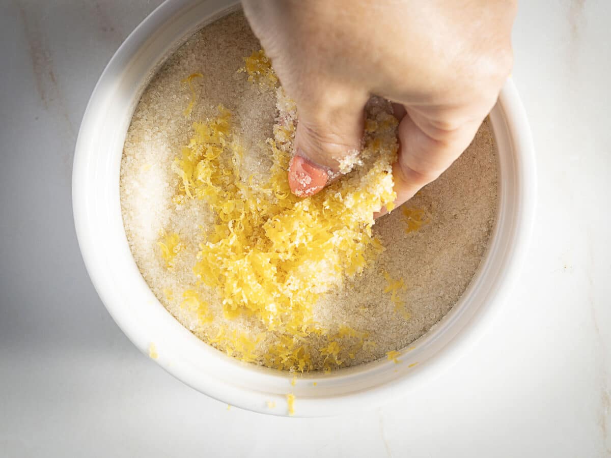 A hand mixes grated lemon zest into a bowl of granulated sugar.