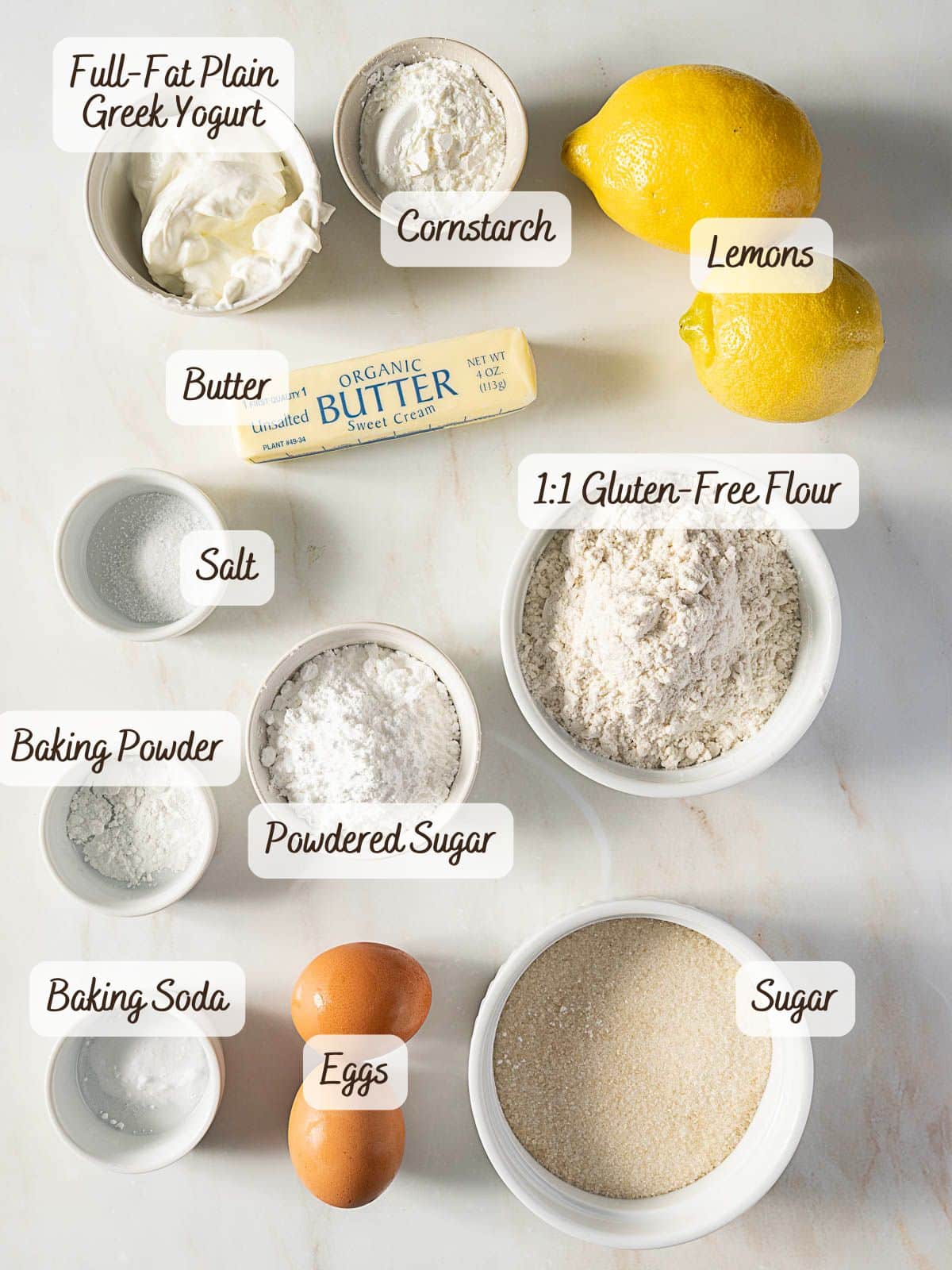 A variety of baking ingredients labeled and arranged on a white surface, ready to create a gluten free lemon pound cake-including eggs, butter, lemons, flour, yogurt, sugar, powdered sugar, cornstarch, salt, baking powder, and baking soda.