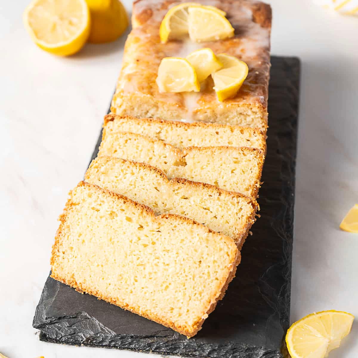 A loaf of lemon bread partially sliced on a slate board, topped with lemon glaze and lemon wedges, surrounded by lemon pieces.