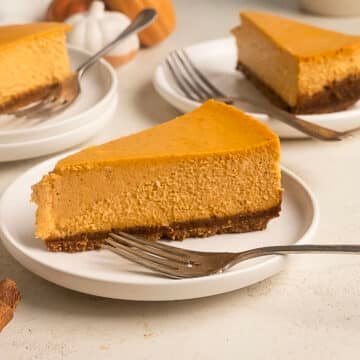 A slice of pumpkin cheesecake with a brown crust sits on a white plate with a fork; additional slices are visible in the background.