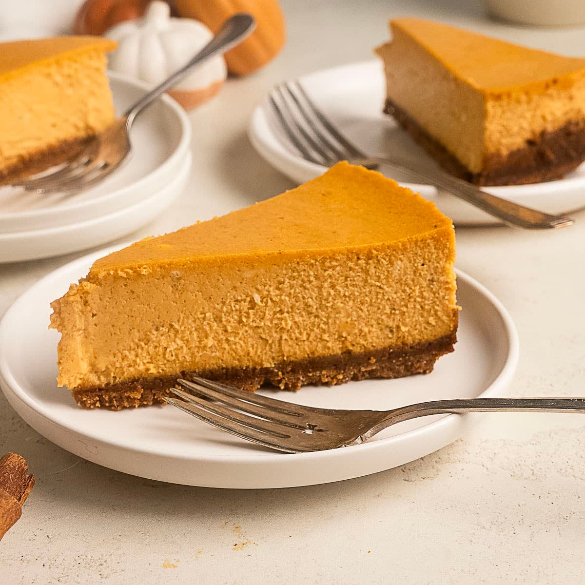 A slice of pumpkin cheesecake with a brown crust sits on a white plate with a fork; additional slices are visible in the background.