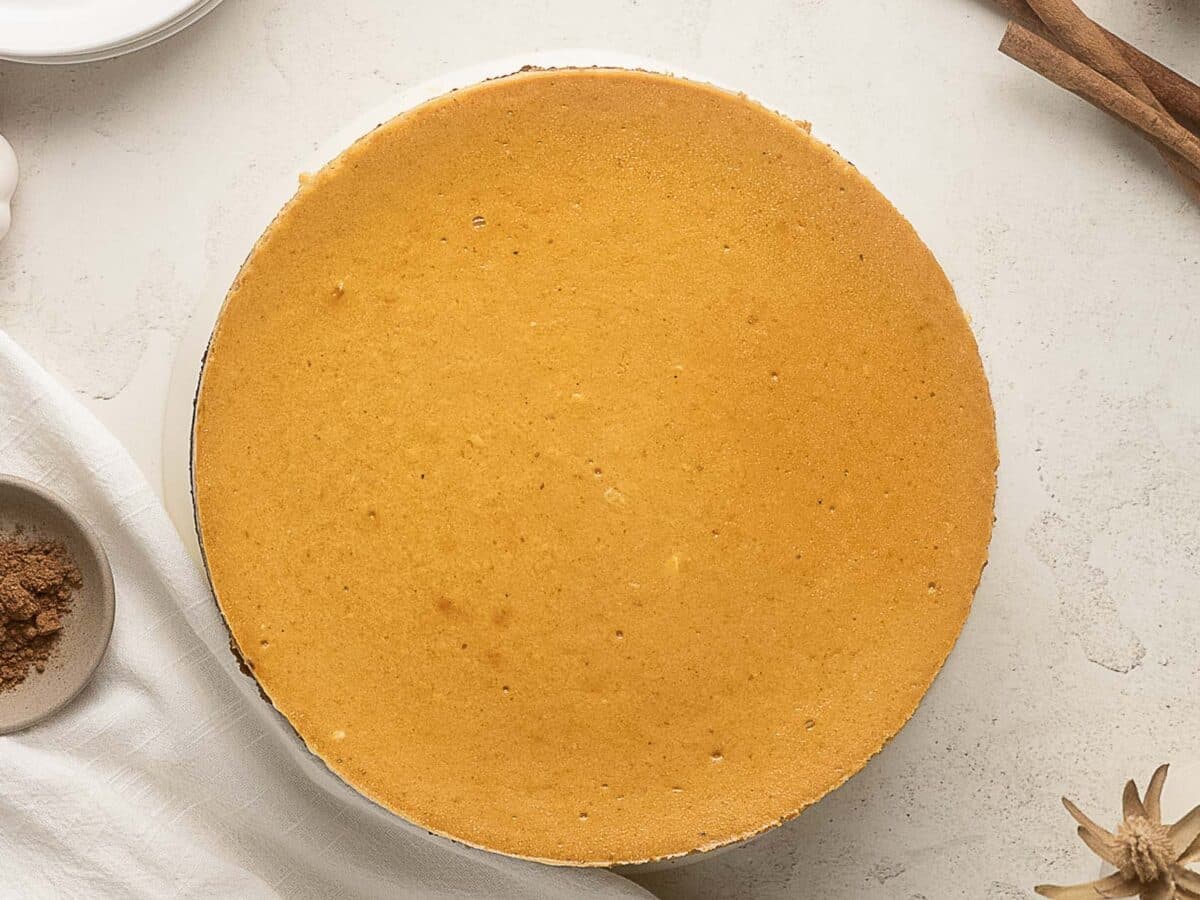 A plain, round pumpkin cheesecake sits on a white surface with minimal decorations and ingredients nearby.