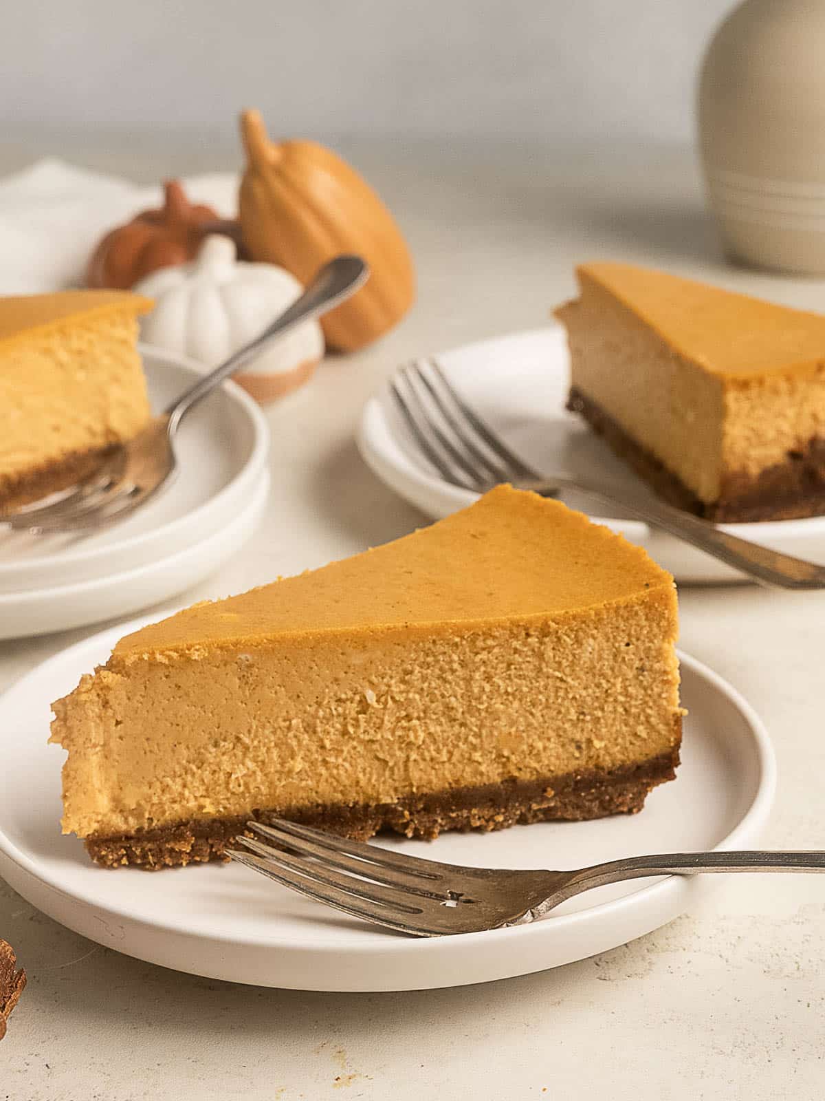 A slice of pumpkin cheesecake with a graham cracker crust on a white plate with a fork, with more slices and decorative gourds in the background.