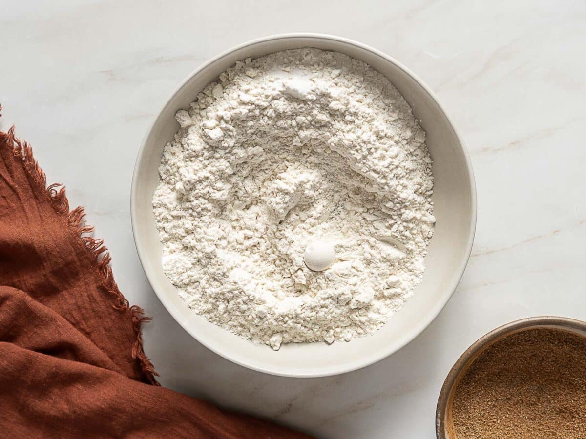 A white bowl filled with flour-perfect for Gluten Free Pumpkin Snickerdoodles-and a pinch of baking powder or salt sits on a light surface next to a brown cloth and a bowl of brown sugar.