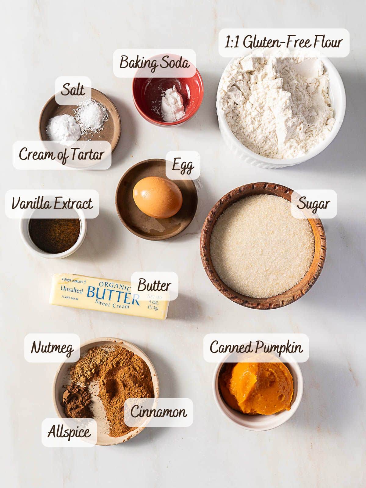 Labeled ingredients for a recipe are arranged on a white surface, including flour, sugar, butter, egg, pumpkin, vanilla, salt, cream of tartar, baking soda, cinnamon, nutmeg, and allspice.