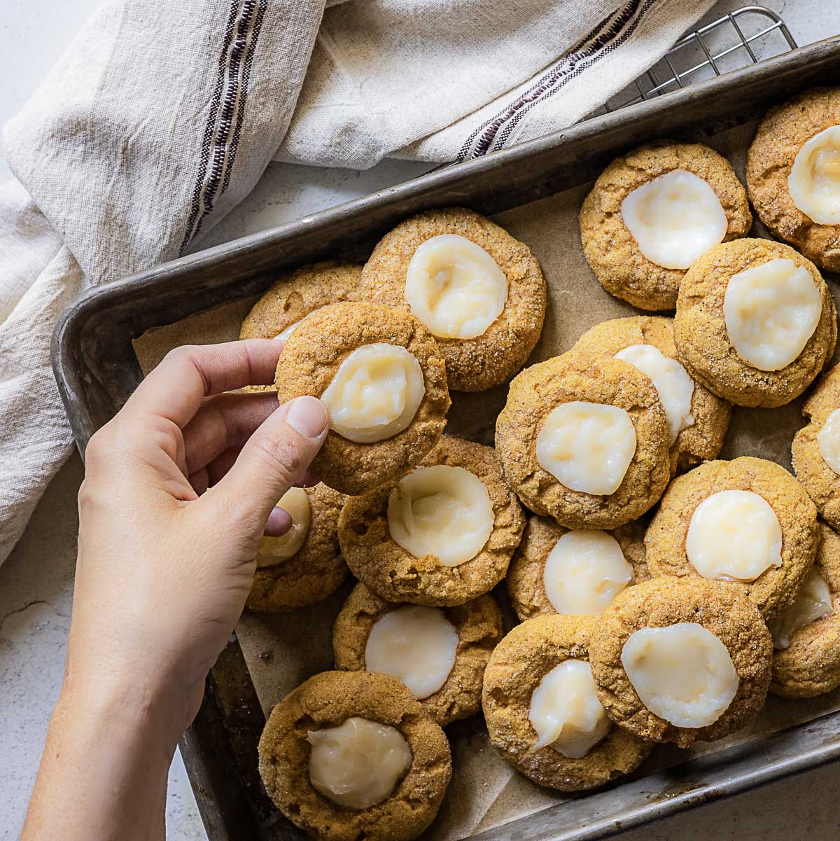 A hand holds a gluten free pumpkin cookie above a baking tray lined with parchment paper and filled with similar cookies. A towel lies nearby.