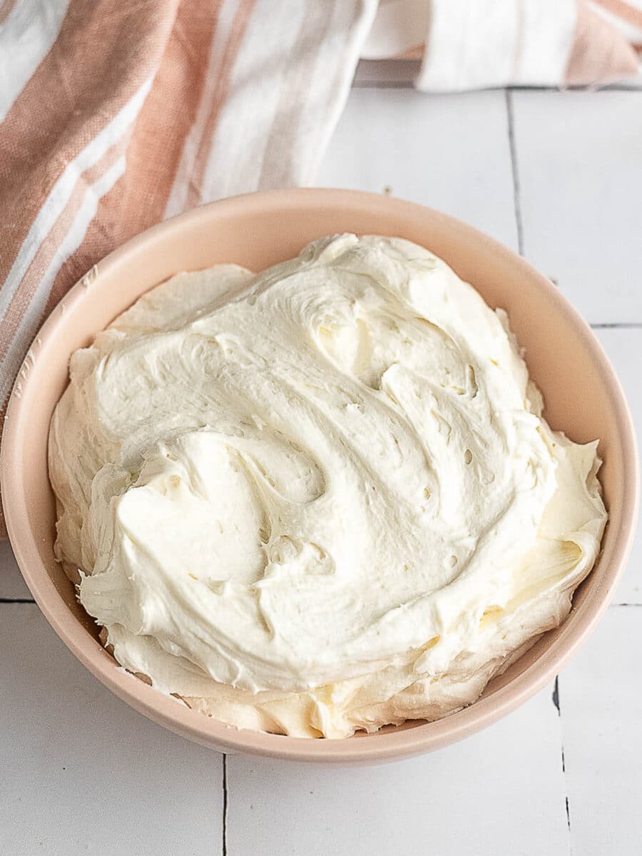 A pink bowl filled with creamy white frosting sits on a white tiled surface next to a pink and white striped cloth.
