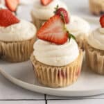 Vanilla cupcakes topped with swirled white frosting and halved fresh strawberries, arranged on a white plate.