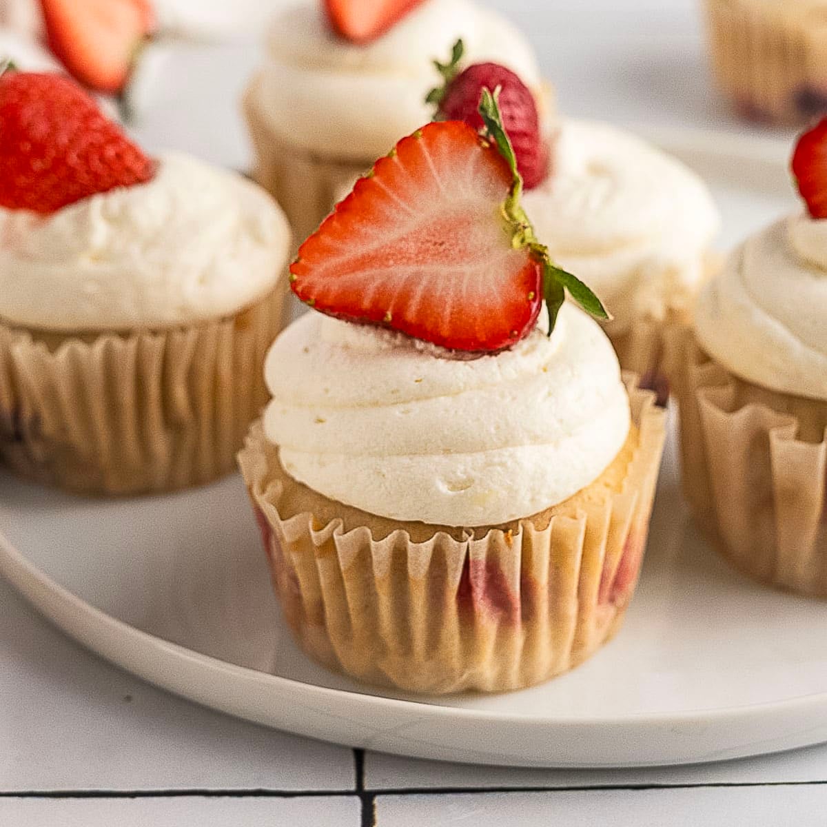 Vanilla cupcakes topped with swirled white frosting and halved fresh strawberries, arranged on a white plate.