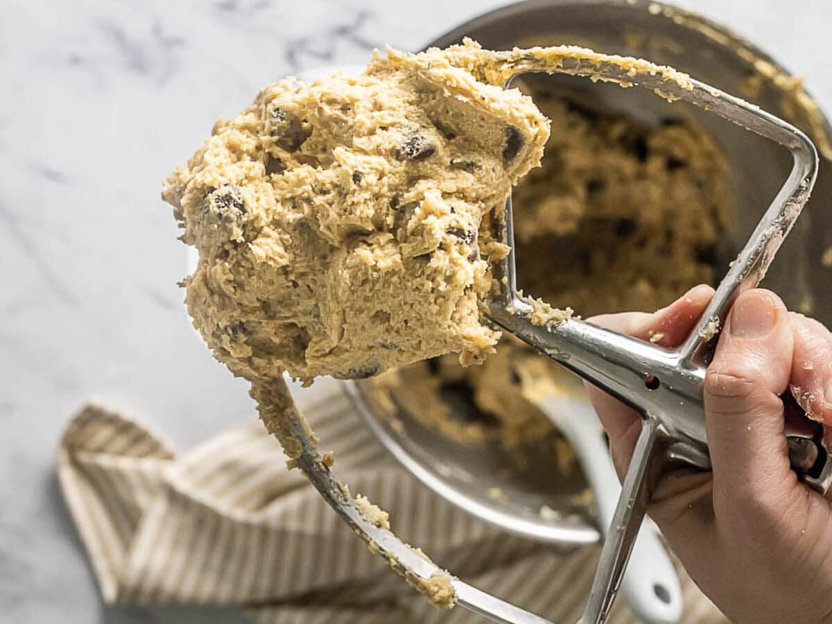 A hand holds a stand mixer beater covered in chocolate chip cookie dough above a mixing bowl on a striped towel.