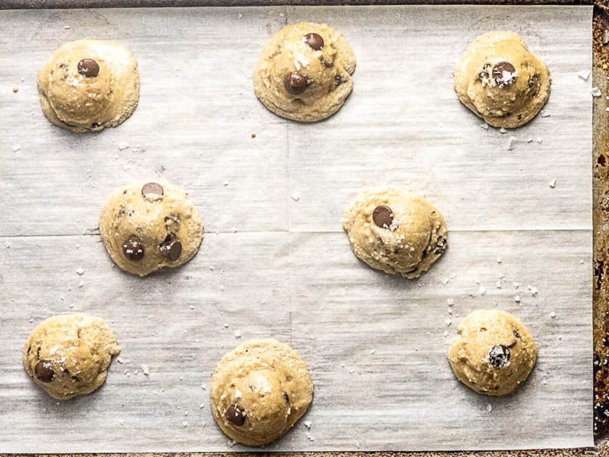 Eight scoops of chocolate chip cookie dough are spaced out on a parchment-lined baking sheet, ready to be baked.