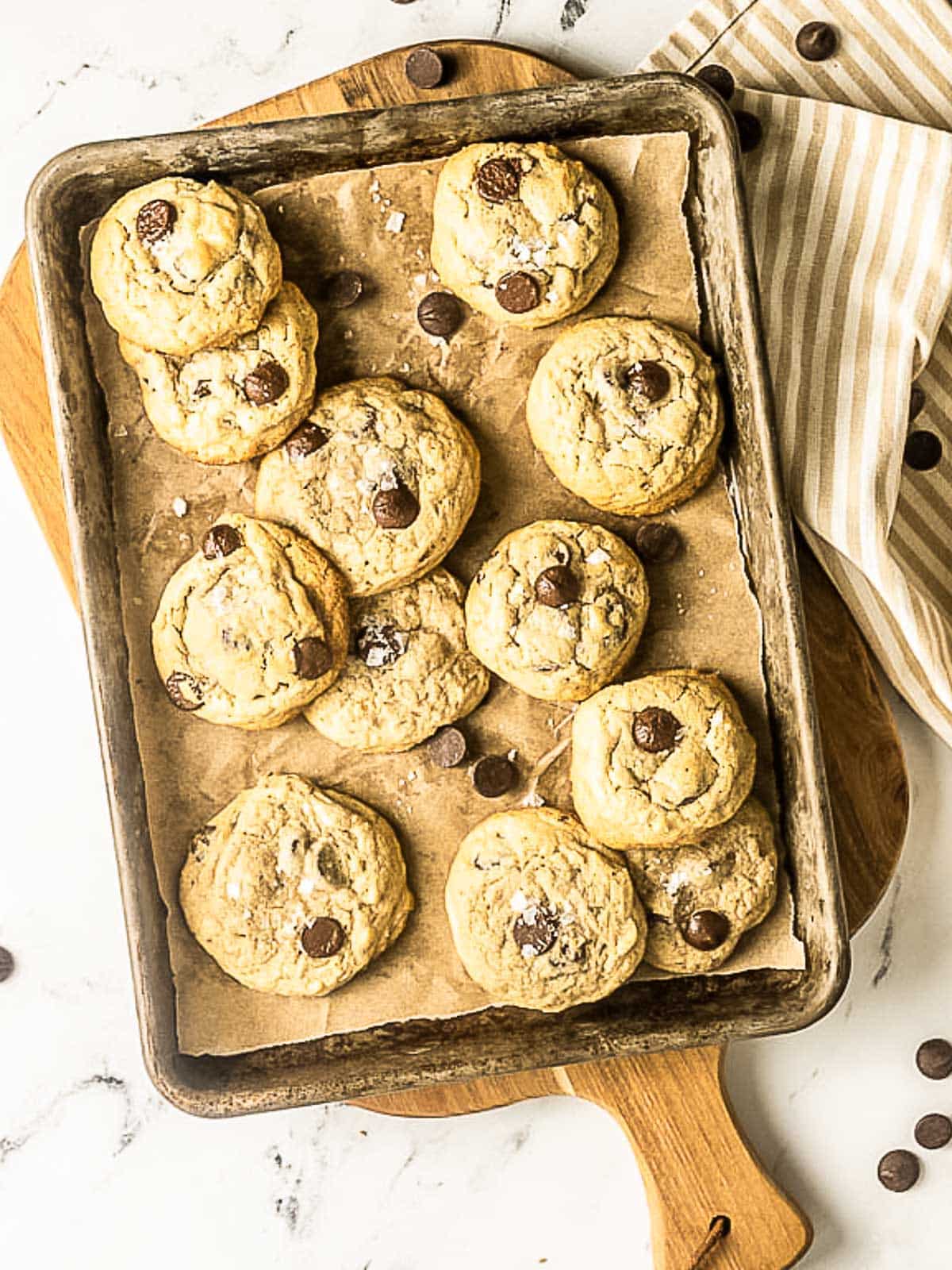 A baking sheet lined with parchment paper holds twelve chocolate chip cookies, some sprinkled with sea salt, on a marble countertop with a striped towel.