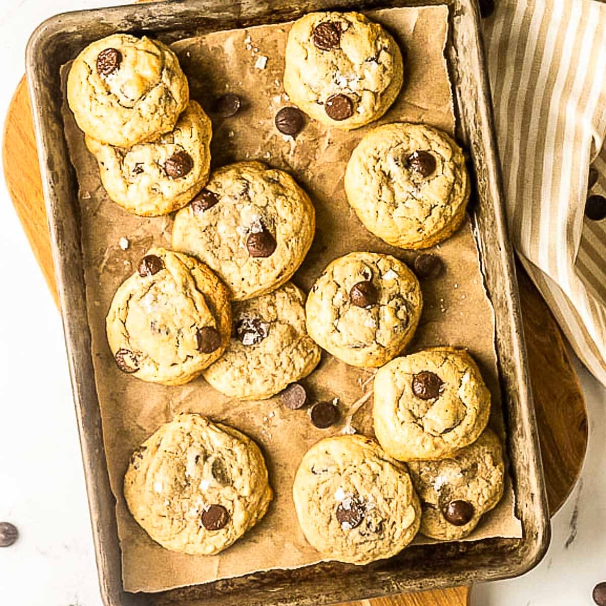 A baking sheet lined with parchment paper holds a dozen chocolate chip cookies, some sprinkled with sea salt, on a light background with a striped napkin nearby.