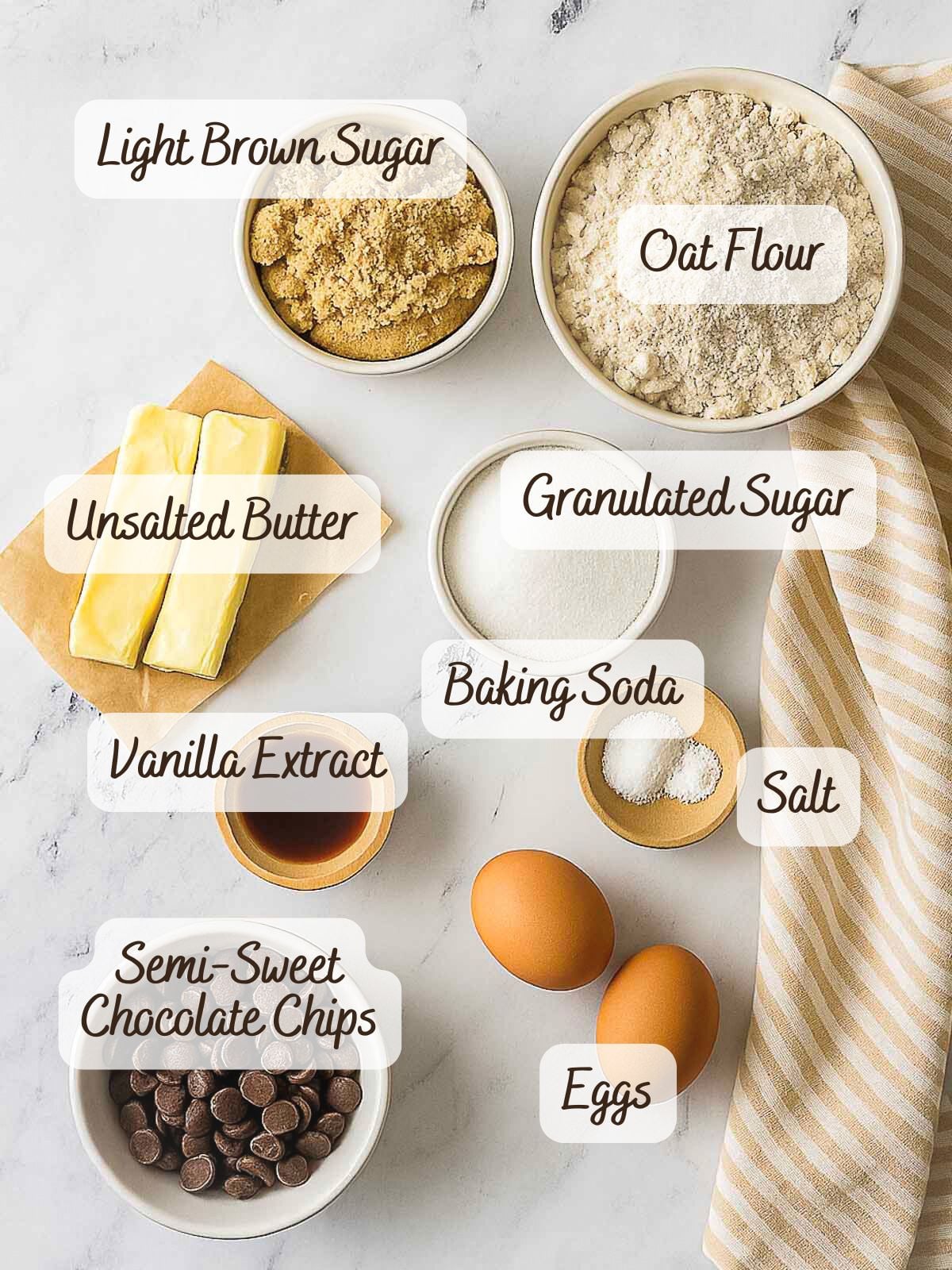 Overhead view of labeled baking ingredients for oat flour chocolate chip cookies: light brown sugar, oat flour, unsalted butter, granulated sugar, vanilla extract, baking soda, salt, semi-sweet chocolate chips, and eggs.