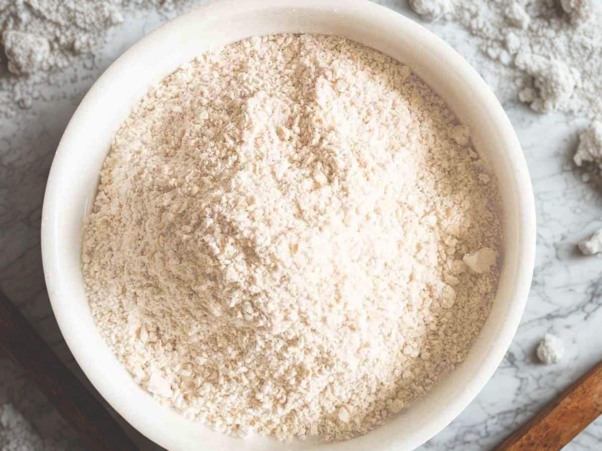 A white bowl filled with a mound of flour sits on a marble surface with scattered flour around it.