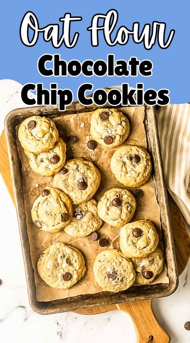 A baking tray lined with parchment paper holds freshly baked oat flour chocolate chip cookies, some sprinkled with sea salt, on a marble surface with a striped cloth nearby.
