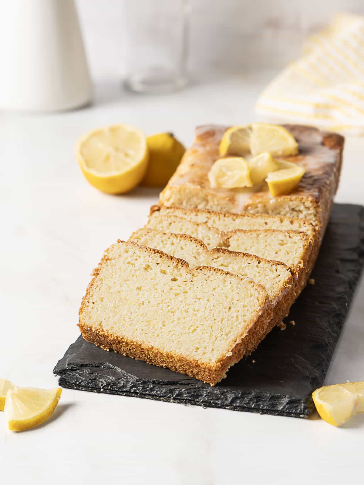 A sliced lemon loaf cake sits on a black tray, topped with lemon slices, with fresh lemon halves and wedges nearby on a white surface.
