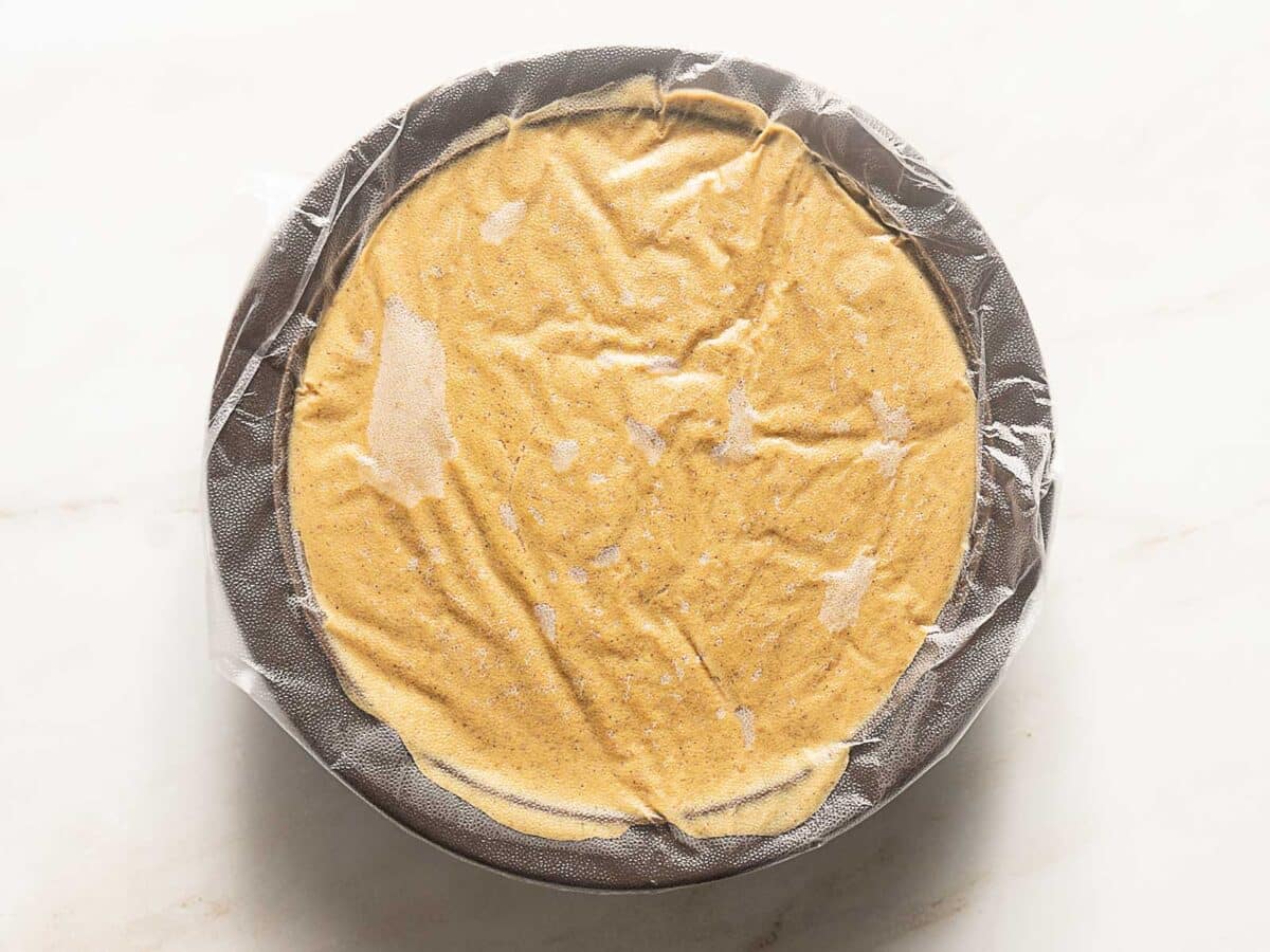 A pie dish filled with an orange batter, covered tightly with plastic wrap, sits on a light-colored surface.
