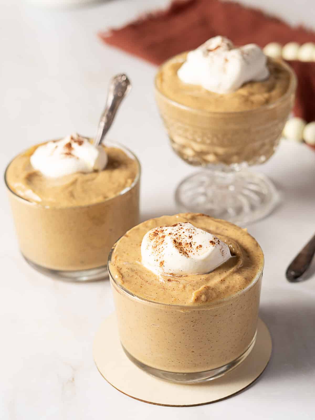Three glasses of pumpkin mousse topped with whipped cream and sprinkled with cinnamon, placed on a white surface with a red cloth in the background.
