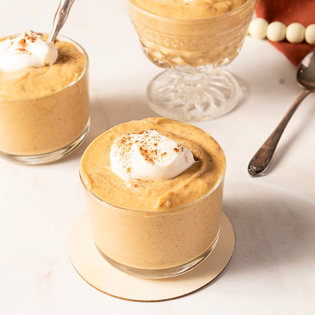 Three glass cups filled with pumpkin mousse, each topped with whipped cream and a sprinkle of spice, placed on a white surface with spoons nearby.