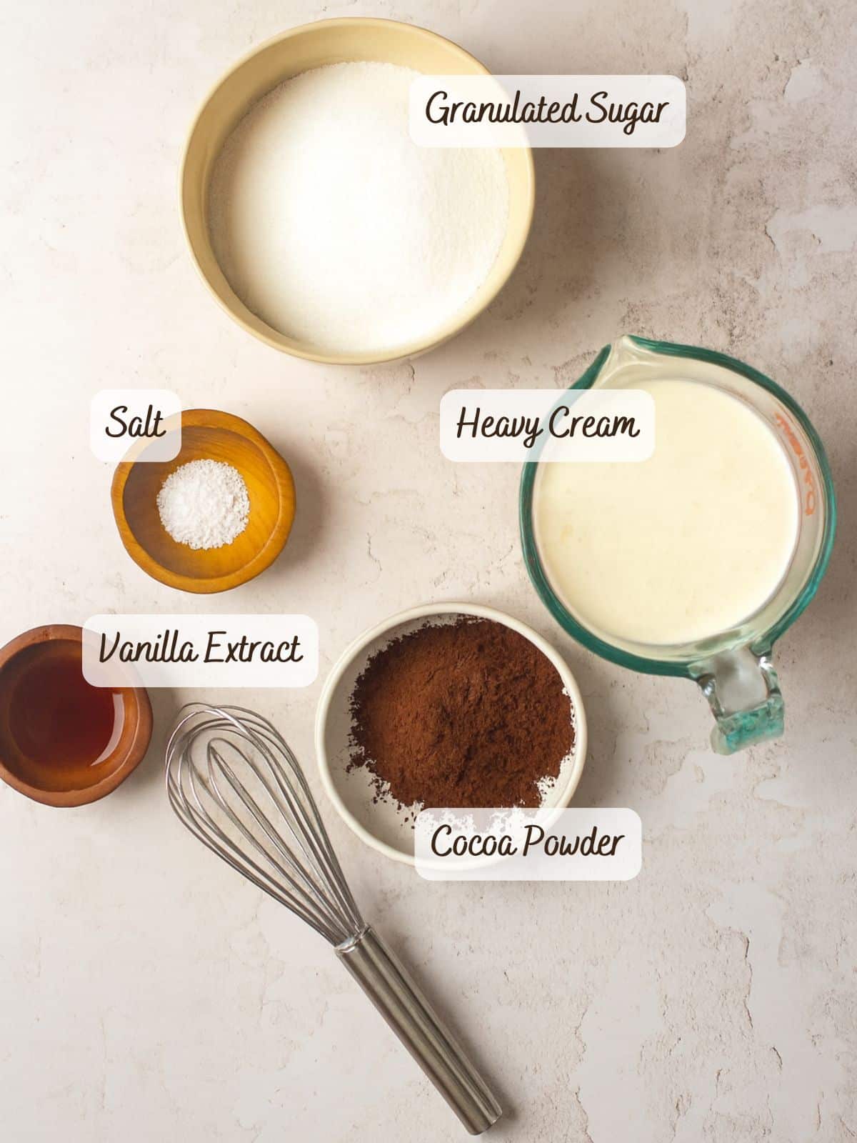 A whisk, granulated sugar, heavy cream, cocoa powder, vanilla extract, salt, and rich chocolate caramel sauce are arranged on a light surface, each labeled with its name.