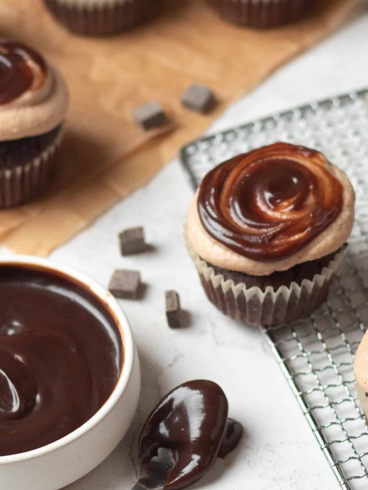 Chocolate cupcakes topped with swirled chocolate ganache sit on a wire rack and parchment paper, with a bowl and spoon of ganache and chocolate chunks nearby.