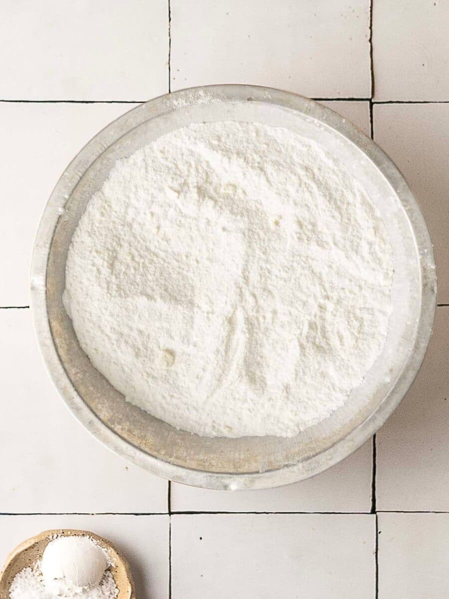 A metal bowl filled with white flour sits on a white tiled surface, with a small bowl of flour visible in the bottom left corner.
