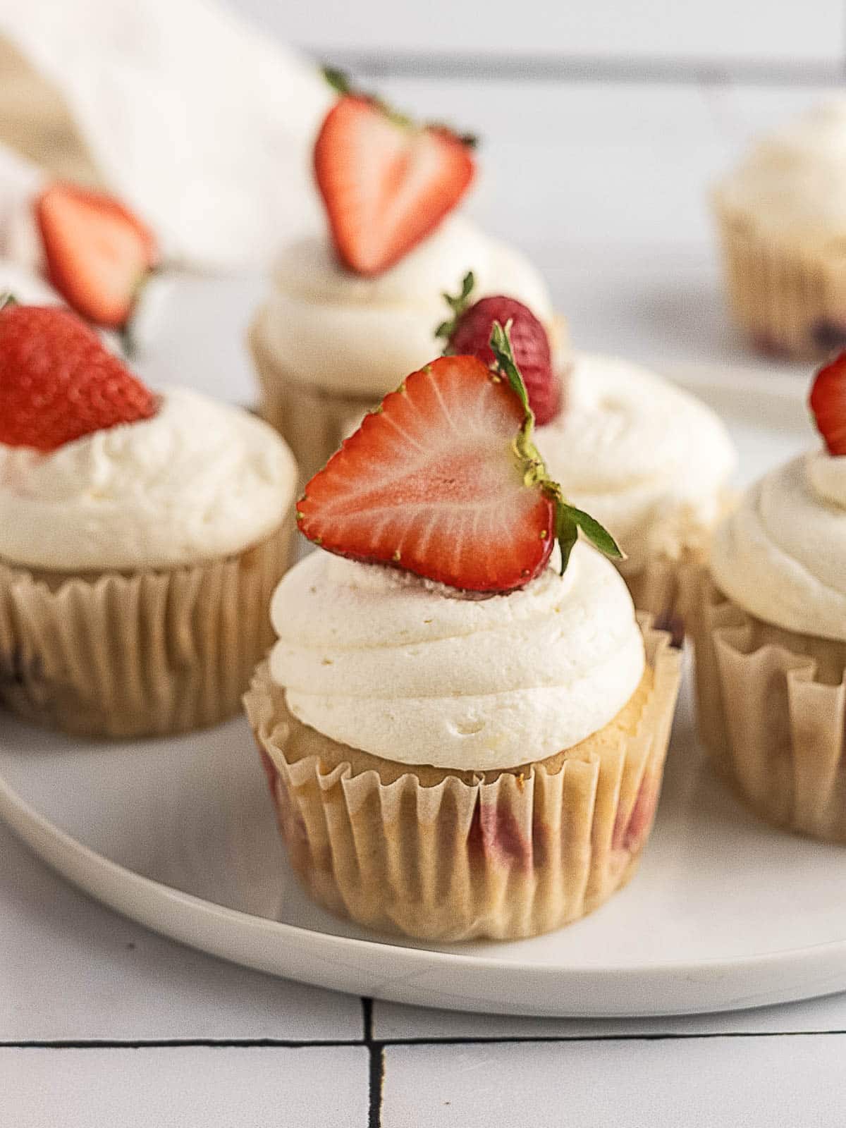 Vanilla cupcakes with white frosting, each topped with a halved fresh strawberry, arranged on a white plate.