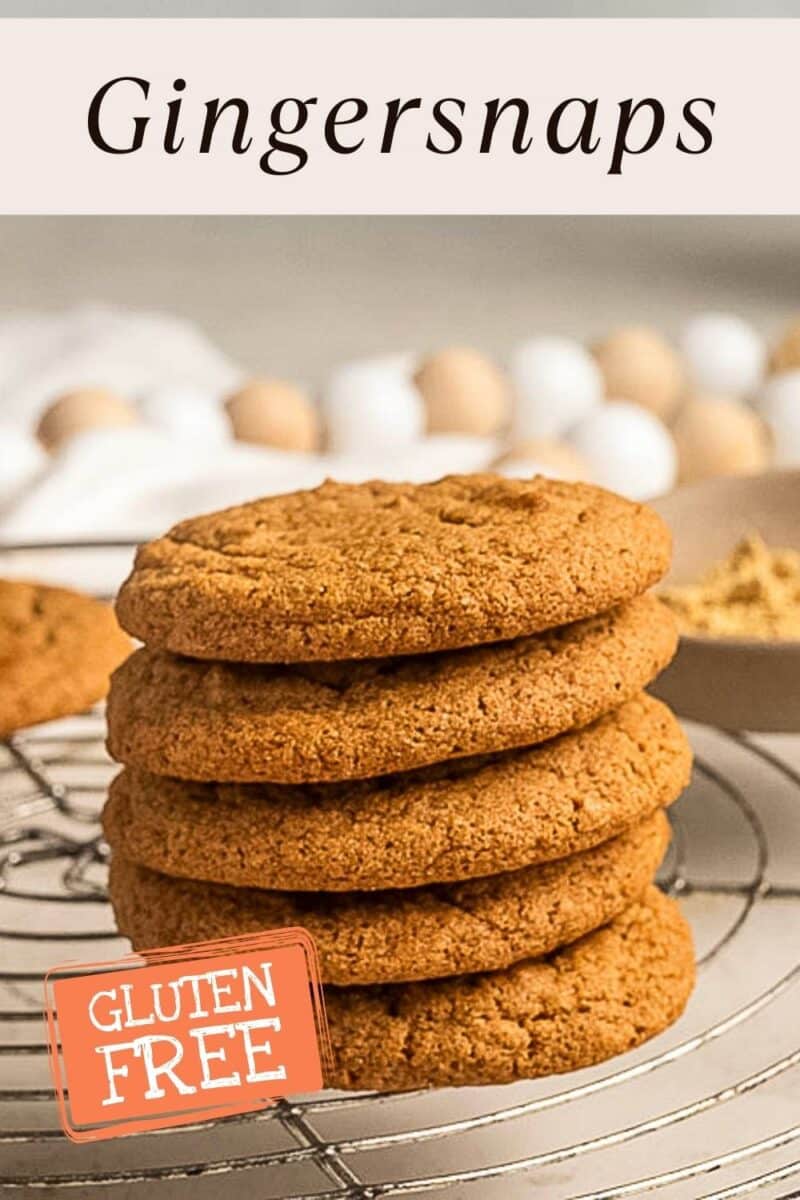 A stack of gluten free ginger snaps rests on a cooling rack with a “gluten free” label; blurred baking ingredients linger in the background.