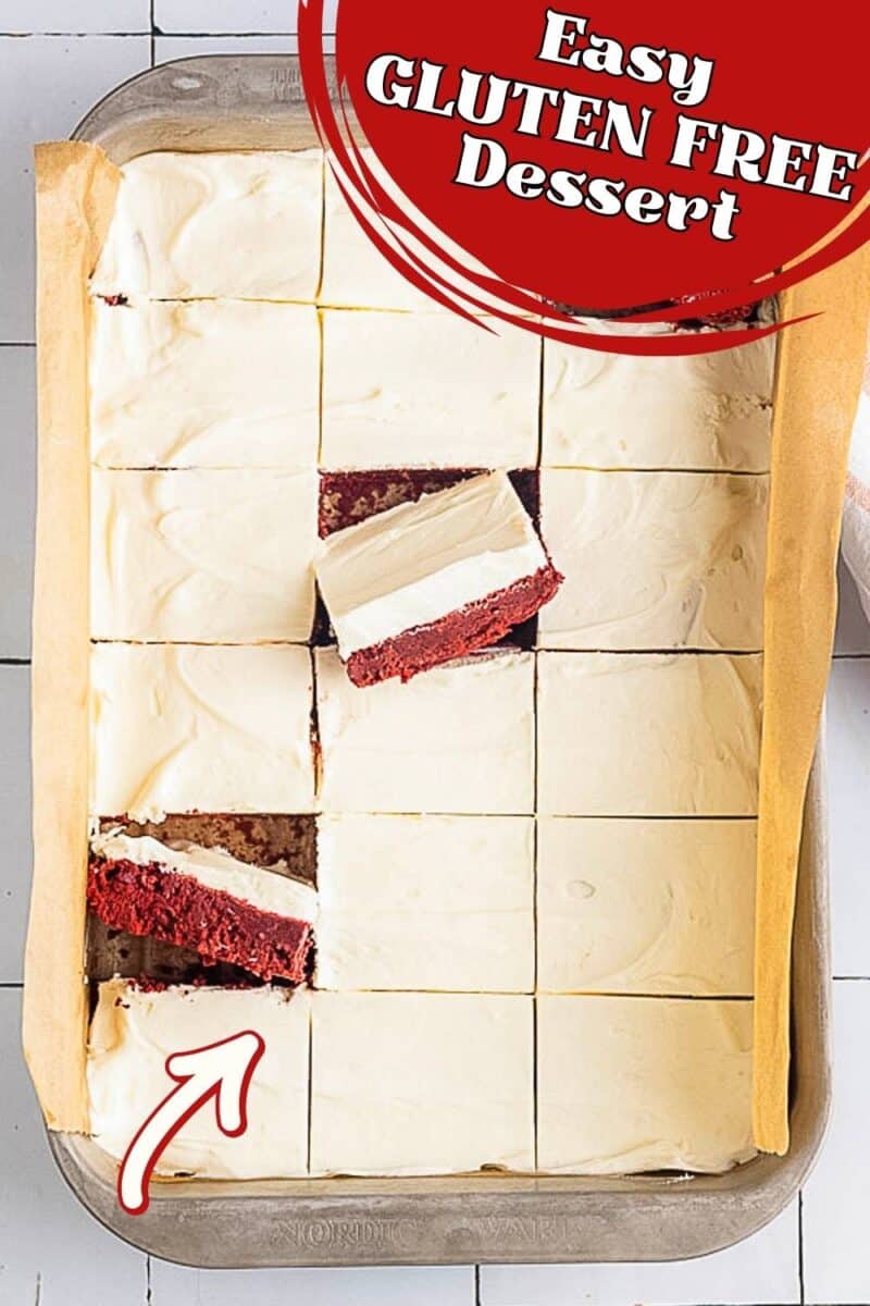 A baking sheet with neatly cut Gluten Free Red Velvet Brownies, each featuring a red velvet layer and white frosting, with one bar pulled out and an arrow pointing to it.