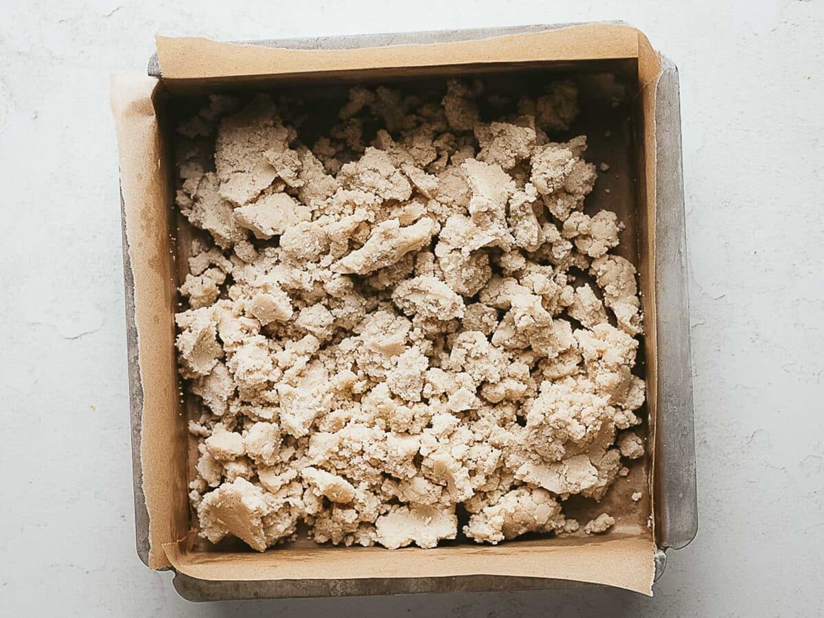A square baking pan lined with parchment paper and filled with an uneven layer of crumbly dough.
