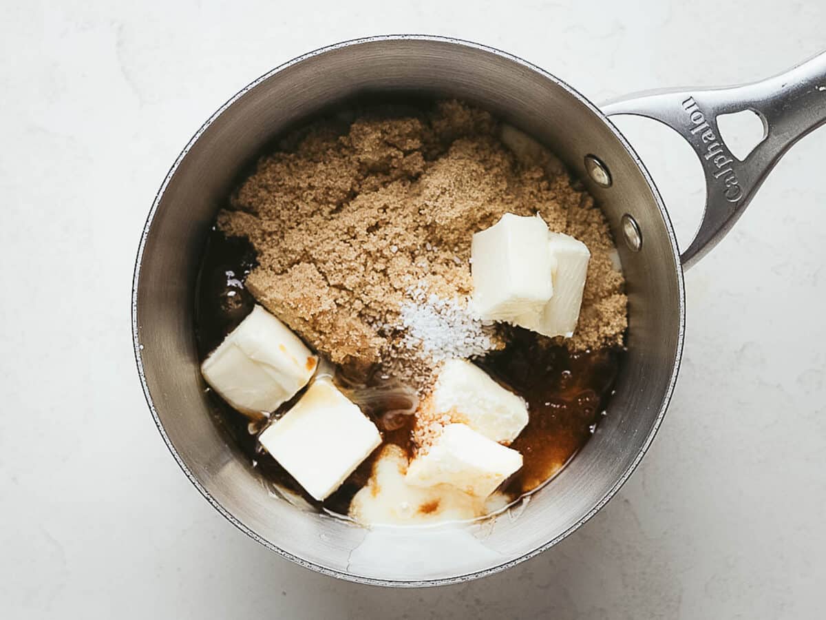 A saucepan containing brown sugar, butter, cream, vanilla extract, and salt on a light surface, ingredients appear unmixed.