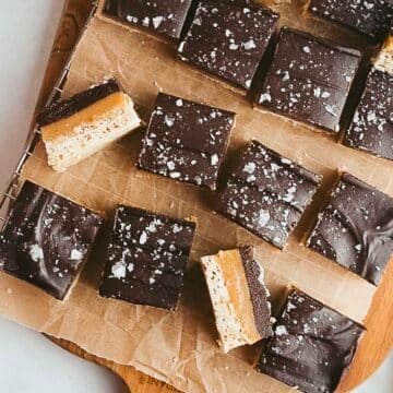 Squares of layered chocolate, caramel, and nougat with a sprinkle of sea salt are arranged on parchment paper on a wooden board.